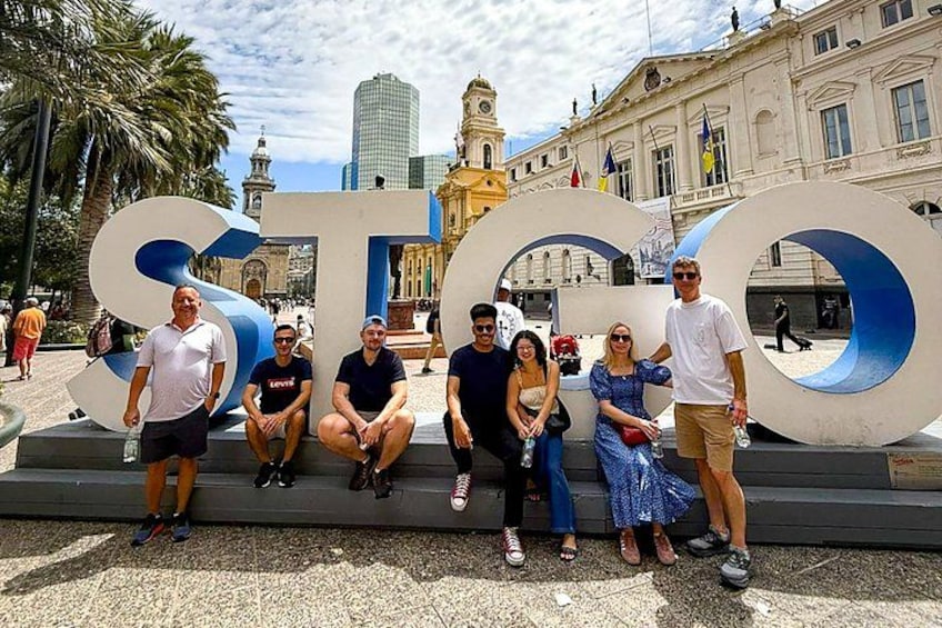 A group of people posing in front of a large, circular sculpture with the number '50' on a plaza with historical buildings in the background. 1
