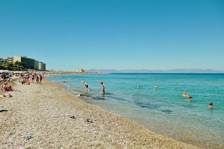 A beach with people swimming and sunbathing, clear blue water, and a distant mountain range. 2
