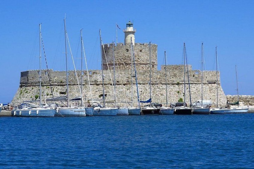 A historic stone fortress with a lighthouse, surrounded by calm blue waters and several sailboats docked at the harbor. 1