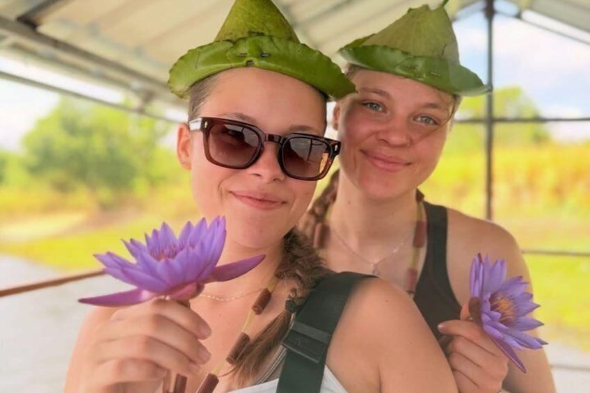 Two people holding purple flowers, wearing hats with leaves, and a structure with a roof in the background. 5