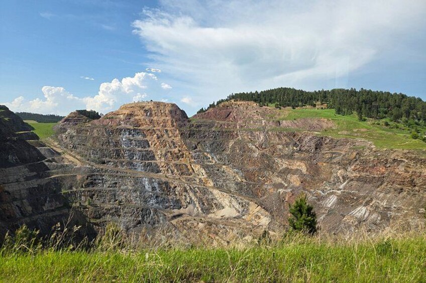 Une vaste mine à ciel ouvert, avec des formations rocheuses en strates et une colline boisée en arrière-plan. 3