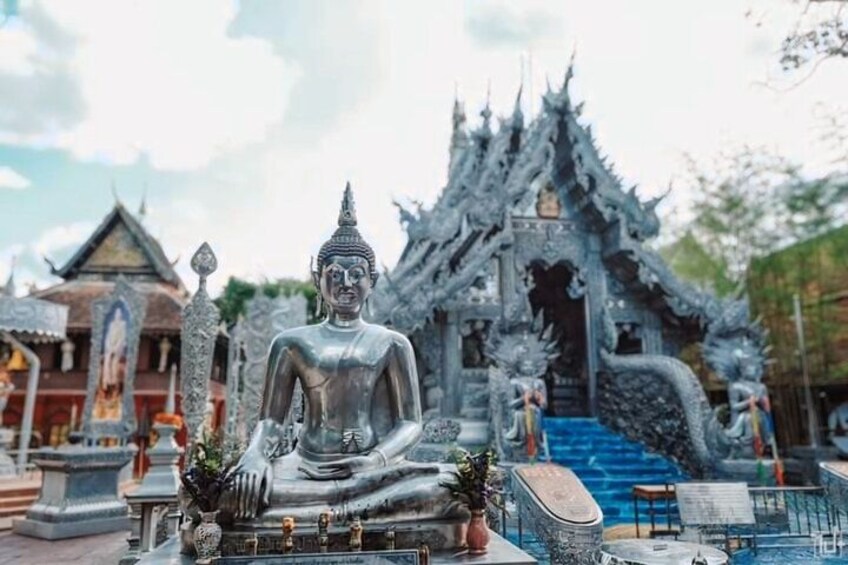 A large seated Buddha statue in front of a traditional Thai temple with ornate architecture. 5