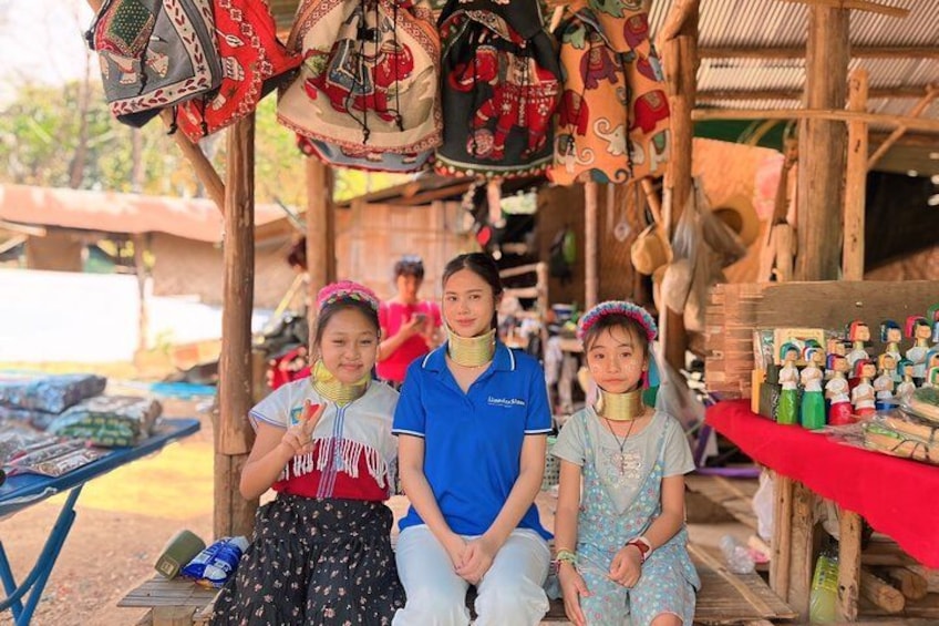 Three individuals seated under a wooden structure with traditional hats and textiles displayed above. 1
