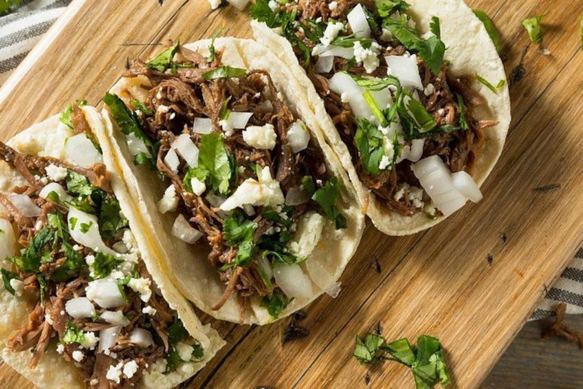 Drei Tacos mit gezupftem Fleisch, gewürfelten Zwiebeln und Koriander auf einem Holzbrett. 2