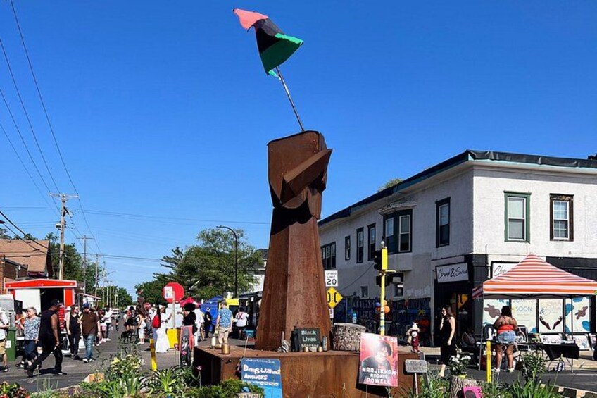 Una plaza pública con una escultura de madera, una calle con peatones y edificios con tiendas. 3