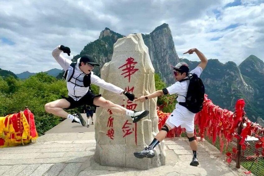 Deux personnes exécutent une figure d’arts martiaux devant un monument de pierre orné d’inscriptions rouges, le tout étant entouré de montagnes et délimité par une barrière faite d’une corde rouge. 1