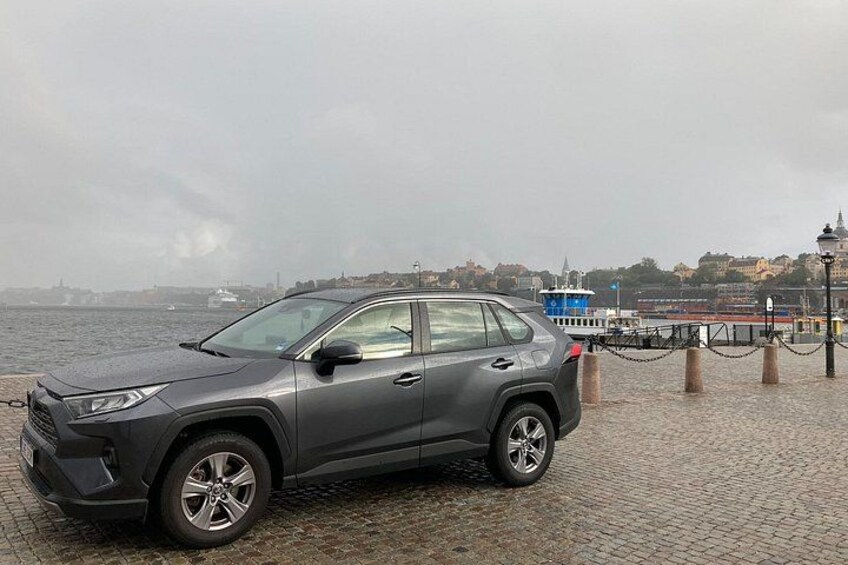 Un SUV gris estacionado en un muelle adoquinado con un barco azul y un paisaje urbano distante al fondo. 3