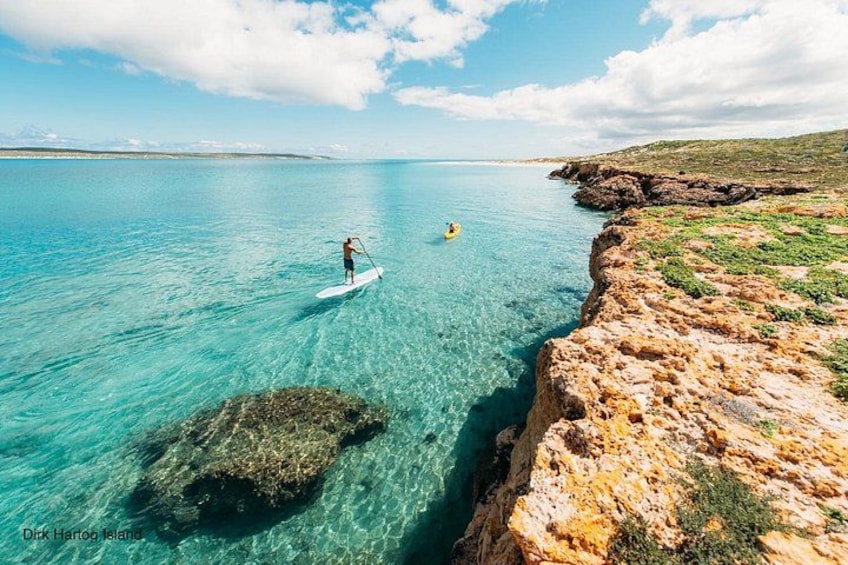 En person som åker stående på en bräda på det klara, turkosfärgade vattnet nära en klippig kust med gles vegetation. 4