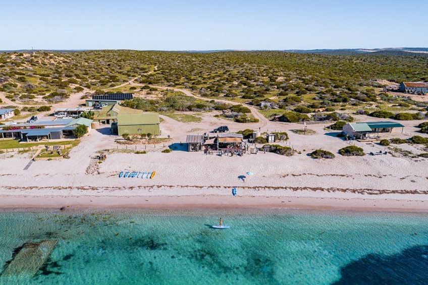 Eine Küstenlandschaft mit Strand, klarem türkisblauem Wasser und einer Siedlung mit Gebäuden und einer Straße. 1