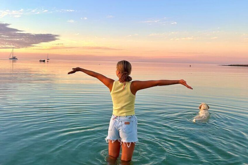 A person standing in shallow water with arms outstretched, facing a sunset over a calm sea with boats in the distance. 4