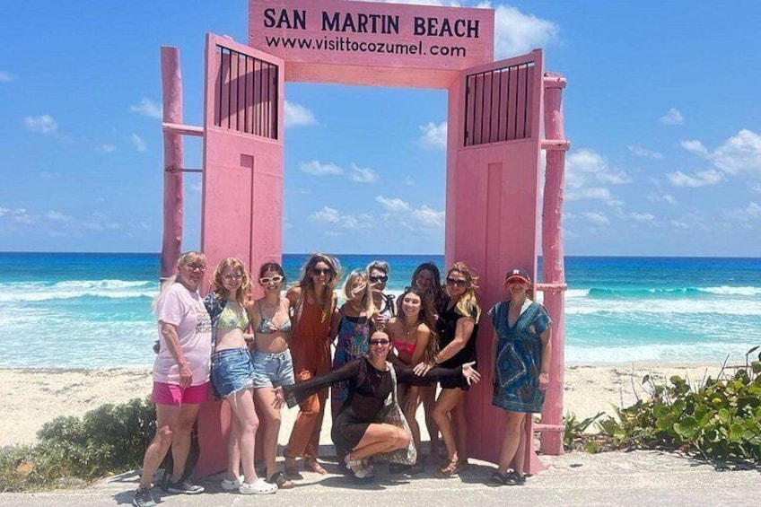 Un grupo de personas posando frente a una puerta rosa en la Playa San Martín, con el océano al fondo. 5