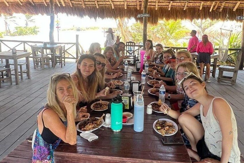 A group of people dining at an outdoor wooden table with a thatched roof, surrounded by tropical vegetation. 3