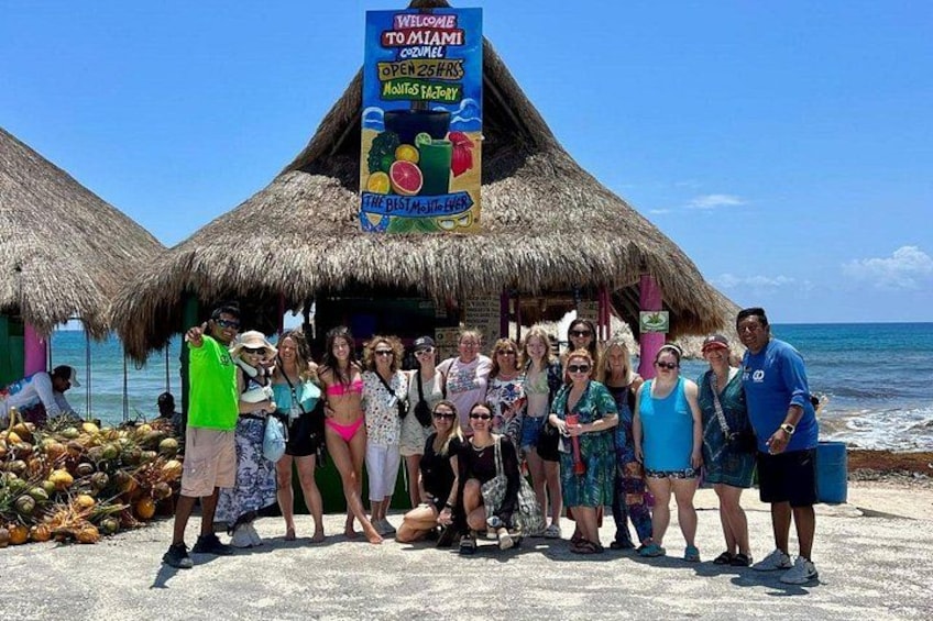 A group of people posing in front of a thatched-roof structure with a signboard advertising local attractions. 4