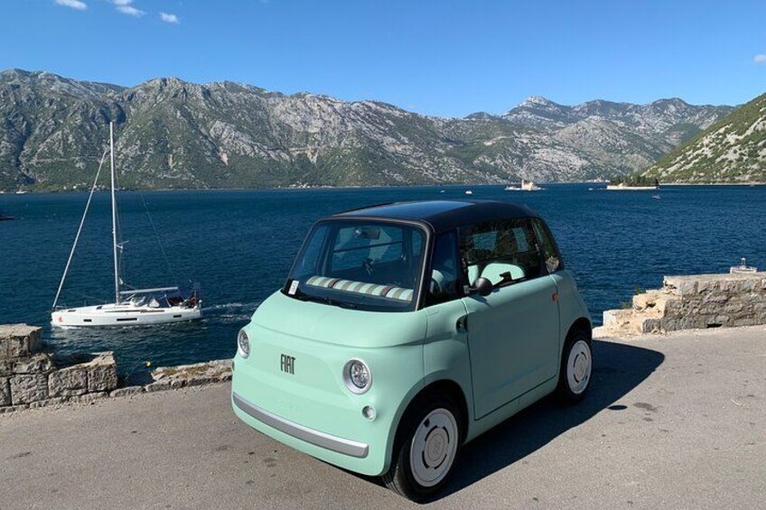 En lysegrøn Fiat cabriolet parkeret ved vandet, med en sejlbåd i baggrunden. 2