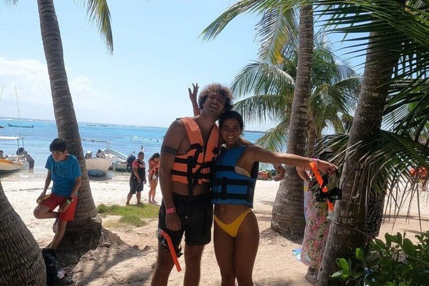 Two people wearing life jackets on a sandy beach with palm trees and boats in the background. 5