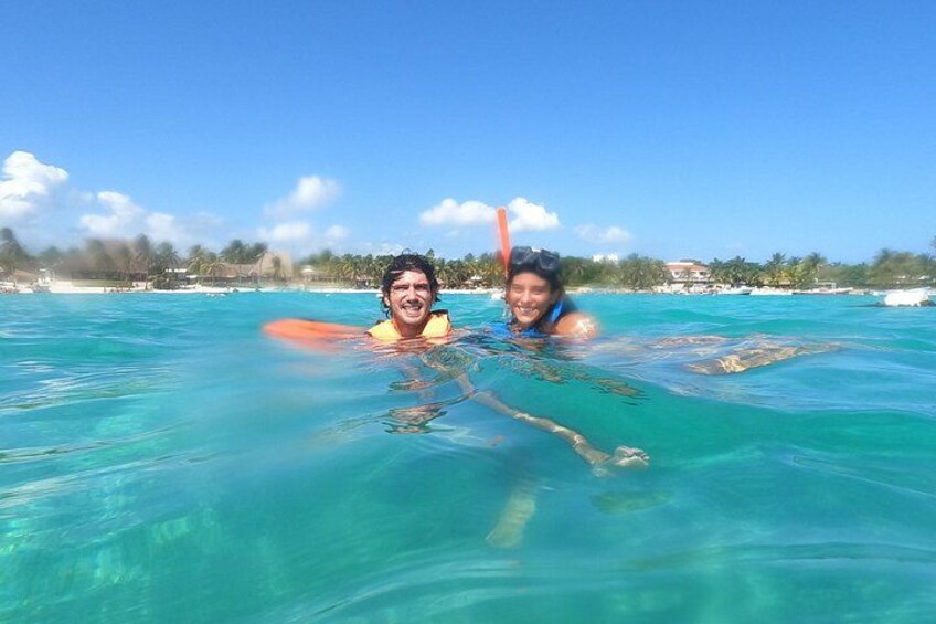 Two people swimming in clear blue water with a beach and palm trees in the background. 4