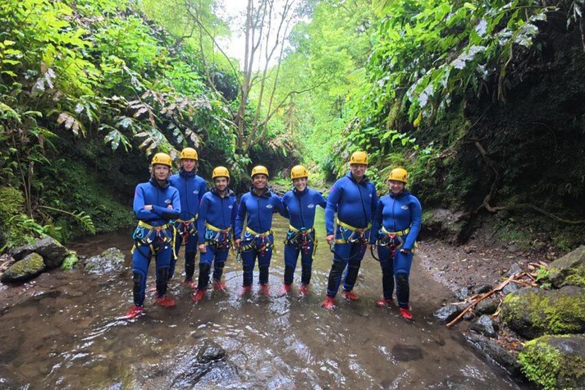A group of people in blue wetsuits and yellow helmets standing in a forested area with a stream. 2