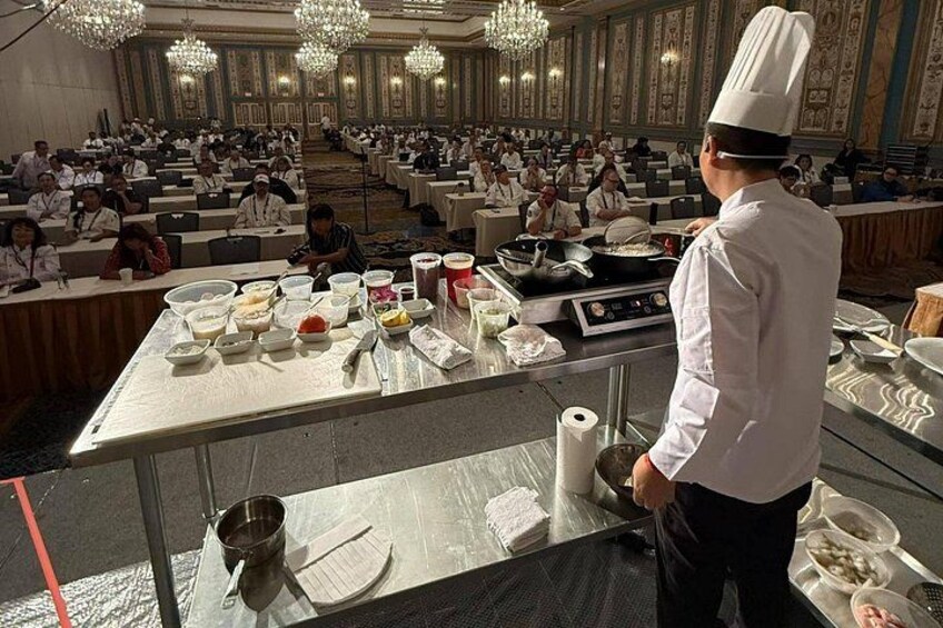 Un chef prépare des plats dans une grande salle de banquet où les participants sont assis à des tables. 2