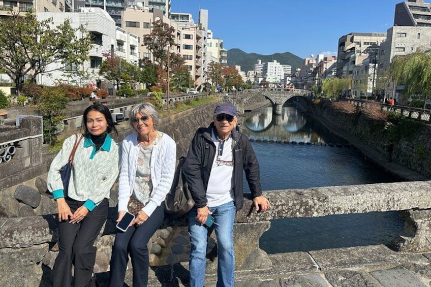 Three people standing by a stone bridge over a river with buildings in the background. 4