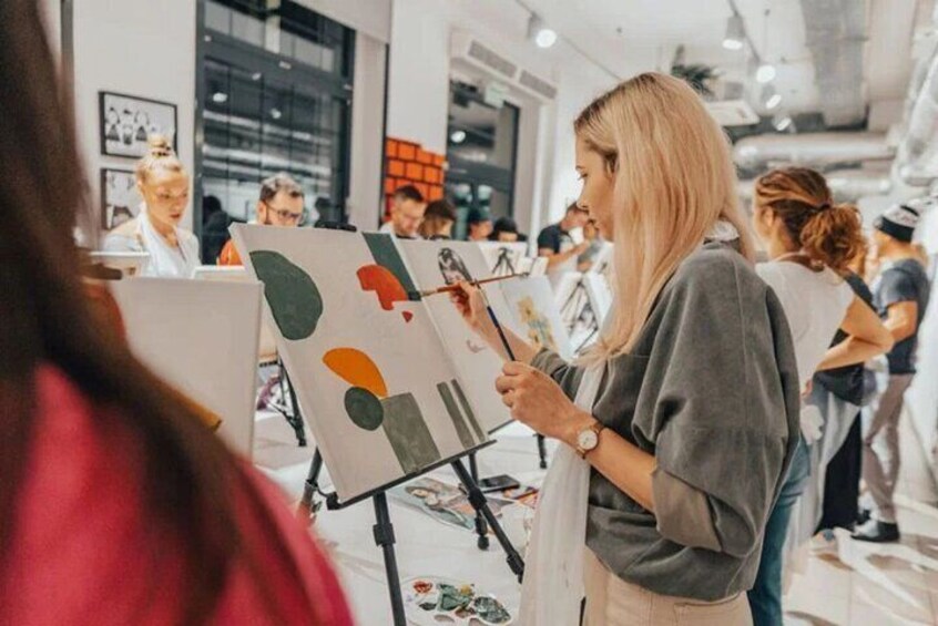 A person painting on an easel in an indoor setting with other people in the background. 4