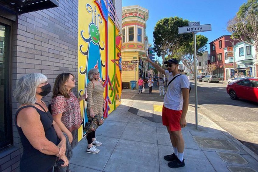 A street scene with a colorful mural, a street sign reading 'Balmy,' and a red car parked on the side. 2