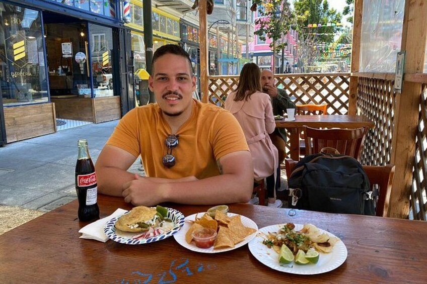 A person sitting at a wooden table with a bottle of Coca-Cola, two plates of food, and a wooden lattice structure in the background. 4