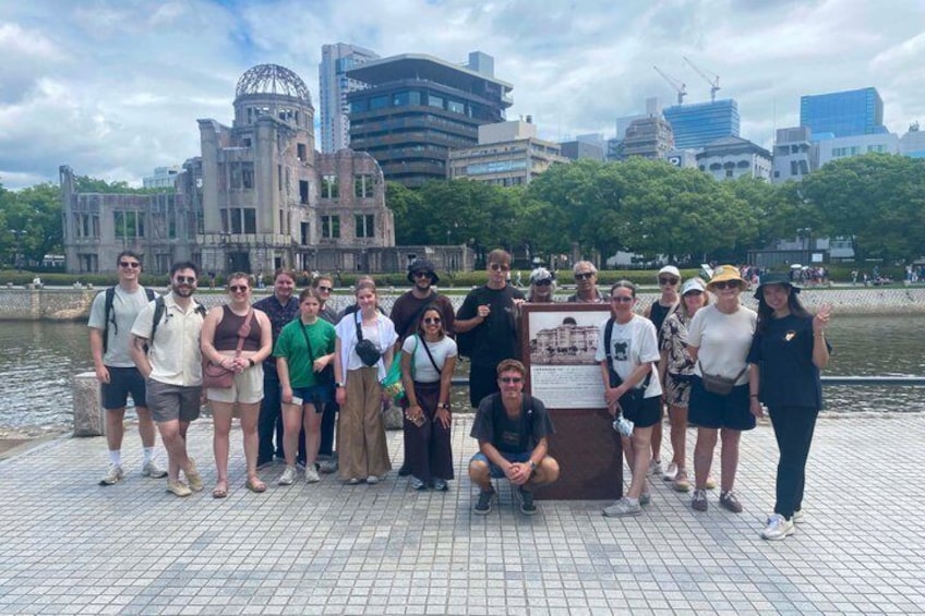 A group of people posing in front of a historical building with a water body and modern skyscrapers in the background. 3