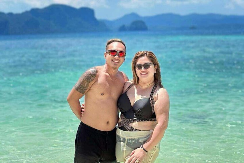 Dos personas de pie en una playa con agua turquesa cristalina y montañas a lo lejos. 4