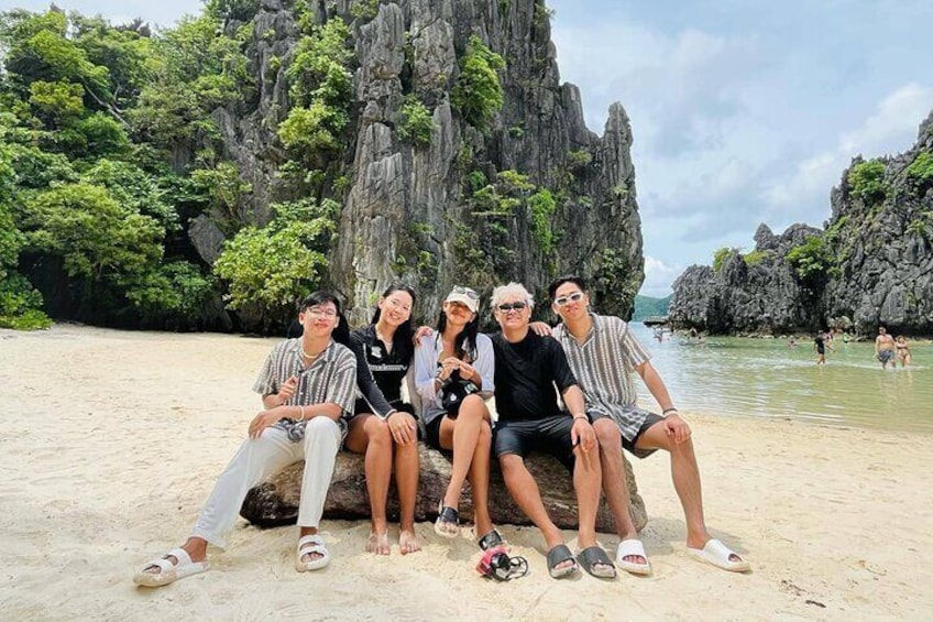 Un grupo de personas sentadas sobre una roca en una playa arenosa, con acantilados rocosos y vegetación al fondo. 3