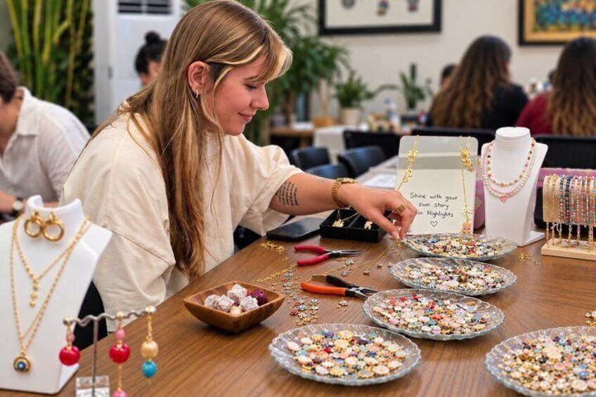 En person laver smykker ved et bord med perler, værktøj til smykkefremstilling og et skilt der siger 'Shine with your Own Style in Highlights'. 1