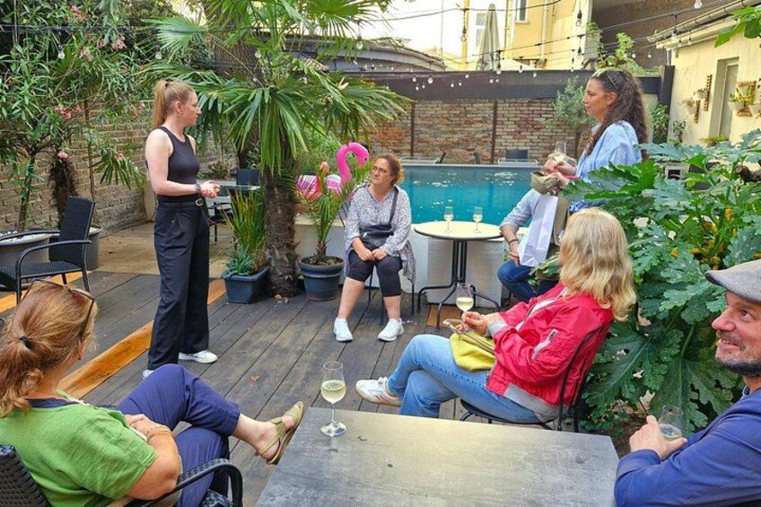 A group of people sitting and standing outdoors, some holding drinks, with a pool and plants in the background. 2