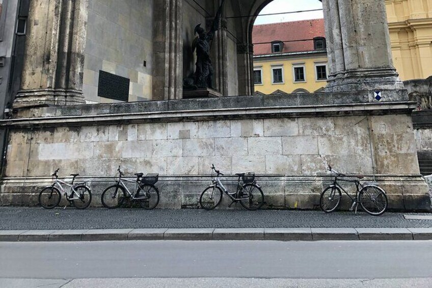 Eine Reihe von Fahrrädern, geparkt an einer Steinmauer, mit einer Statue und architektonischen Säulen im Hintergrund. 4