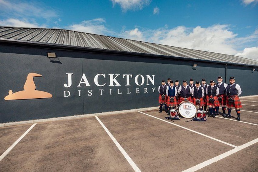 Eine Brennerei namens Jackton, vor der eine Gruppe von Menschen in Kilts und mit Trommeln steht. 5