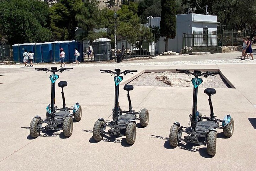 Drie elektrische scooters staan op een betonnen oppervlak in een open gebied met bomen en een gebouw op de achtergrond. 2