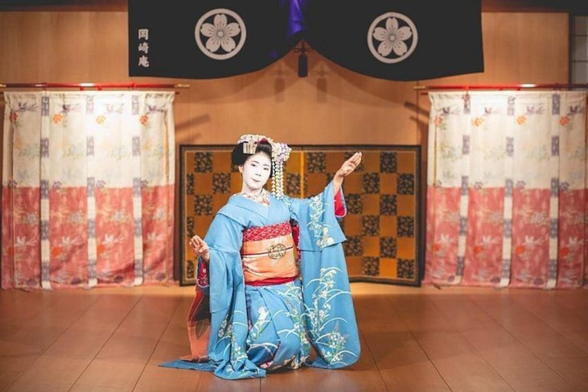 Una persona vestida con traje tradicional japonés actuando en un escenario con un telón de fondo de cortinas estampadas y suelo de madera. 5