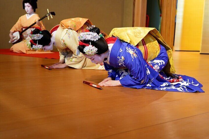 Three performers in traditional Japanese costumes on a wooden floor, one in blue and two in orange, with one holding a fan. 1