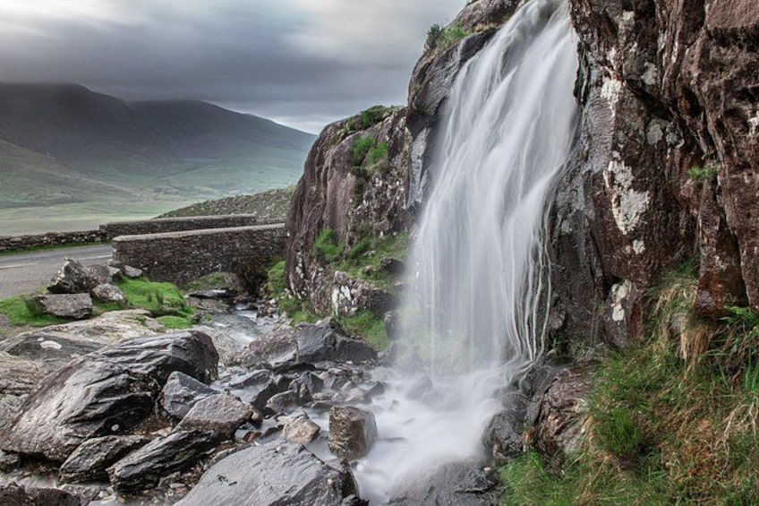 A waterfall cascading down a rocky cliff, surrounded by a grassy landscape and a cloudy sky. 1