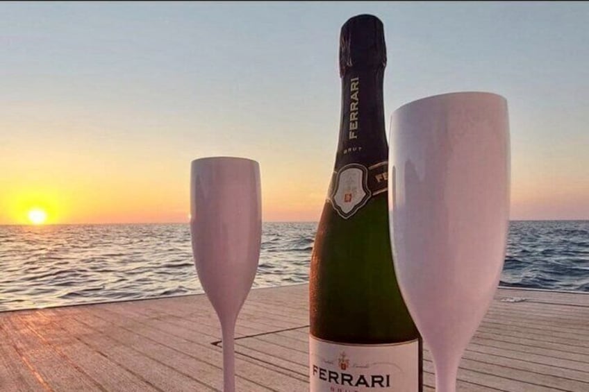 Een zonsondergang boven de oceaan, met een fles Ferrari-wijn en twee glazen op een houten terras. 2