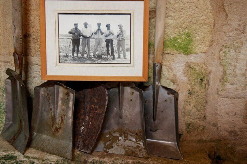 A framed black and white photo of four men standing in a field, mounted on rusted metal plows against a stone wall. 4