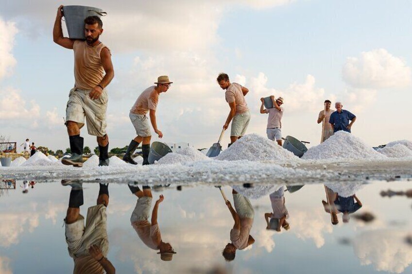People working on a salt farm with large piles of salt and a reflective water surface. 1