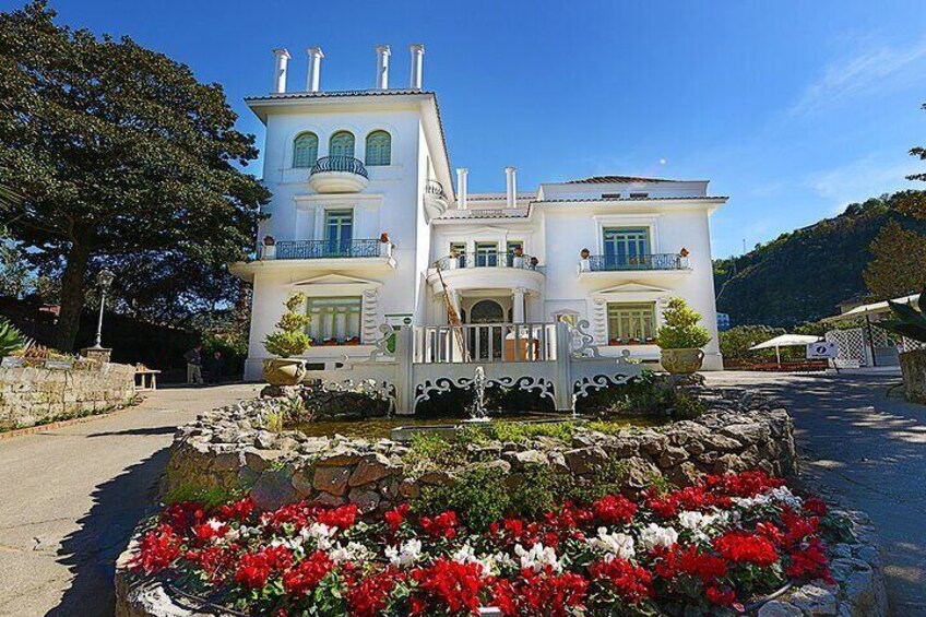 A large white house with multiple balconies and a garden with red and white flowers. 1