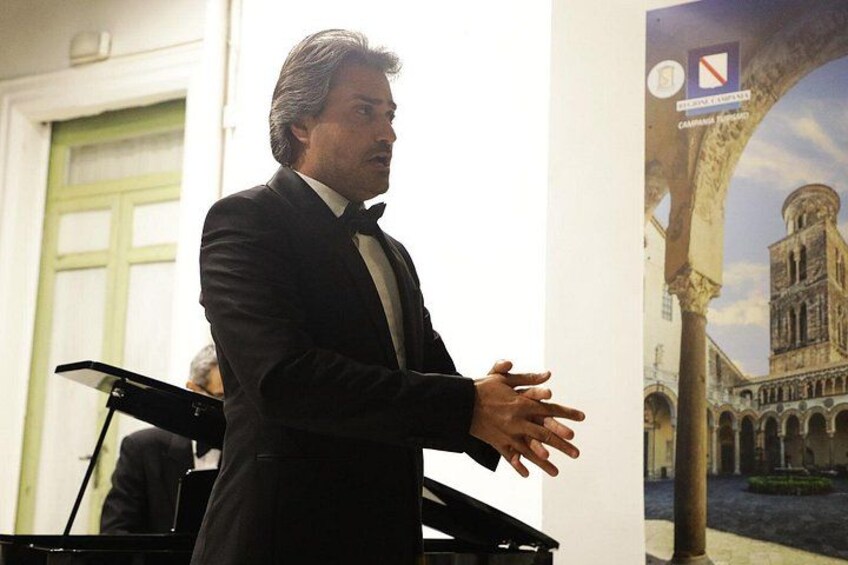A man in a black suit speaking at a podium with a poster of a historical building in the background. 2
