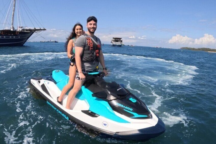 Two people on a jet ski in the ocean with a sailboat and a small island in the background. 5