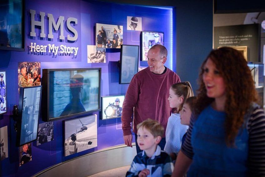 A museum exhibit with a blue wall and various displays, including a large screen and several framed pictures. 2