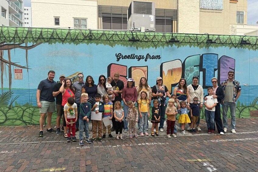 A group of people standing in front of a mural with the text 'Greetings from' and a partial view of a cityscape in the background. 3
