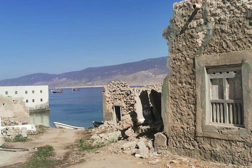 Eine Küstenlandschaft mit einem Steinhaus, einer teilweise baufälligen Struktur und Booten auf dem Wasser. 1