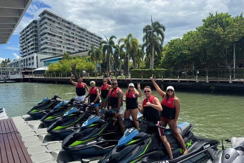Een groep mensen op jetski's aan een waterkant, met palmbomen en een gebouw op de achtergrond. 4