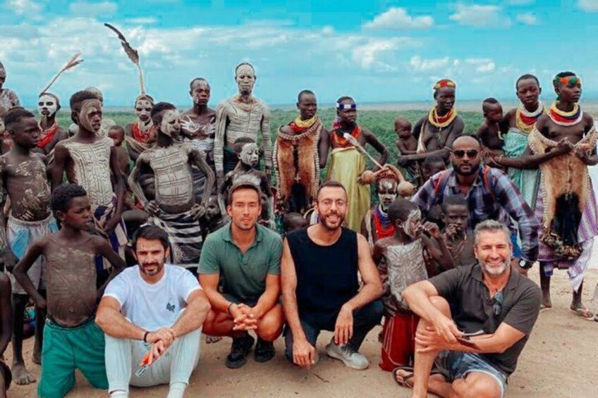A group of people in traditional attire and modern clothing, standing and sitting on a dirt ground. 3