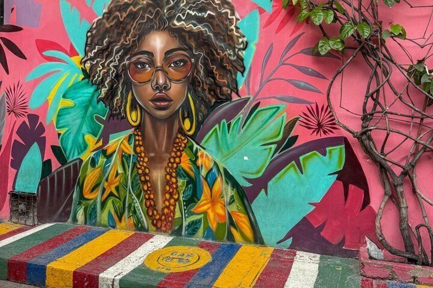Un mural con una persona vestida con un atuendo colorido y un collar, rodeada de vibrantes hojas y plantas tropicales. 5