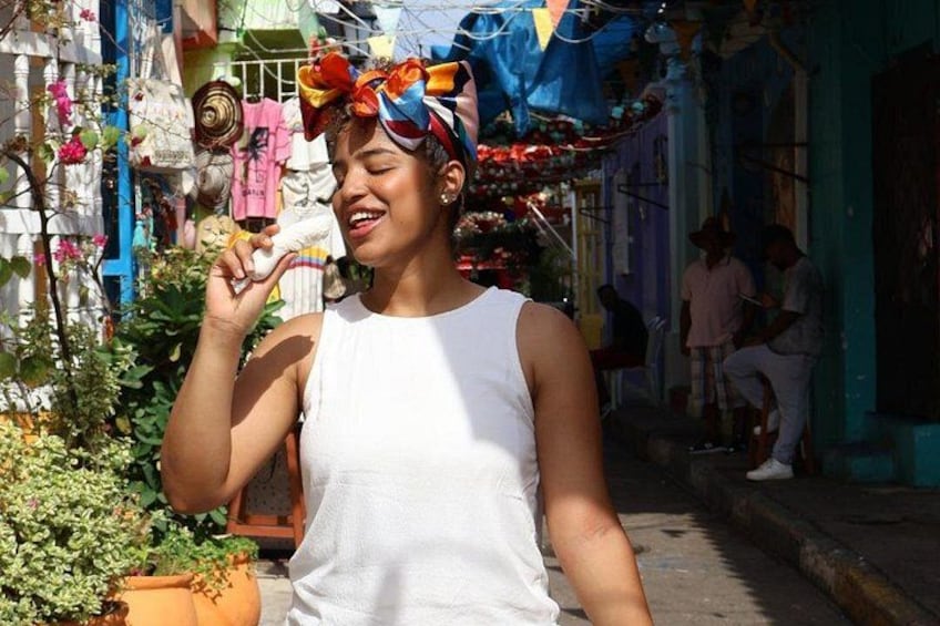 A person in a white tank top and colorful headscarf walking on a street with potted plants and buildings in the background. 1